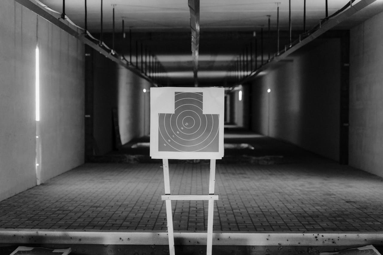 Black and white photo of a shooting range with a focus on a paper target in a dimly lit indoor setting.